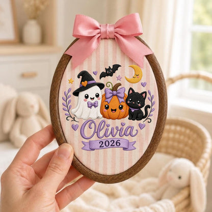 May include: An oval wooden embroidery hoop featuring a light pink and white striped fabric background. The design includes a ghost, a pumpkin, and a black cat, with the name Olivia and the year 2026 printed below. A pink satin bow sits on top.