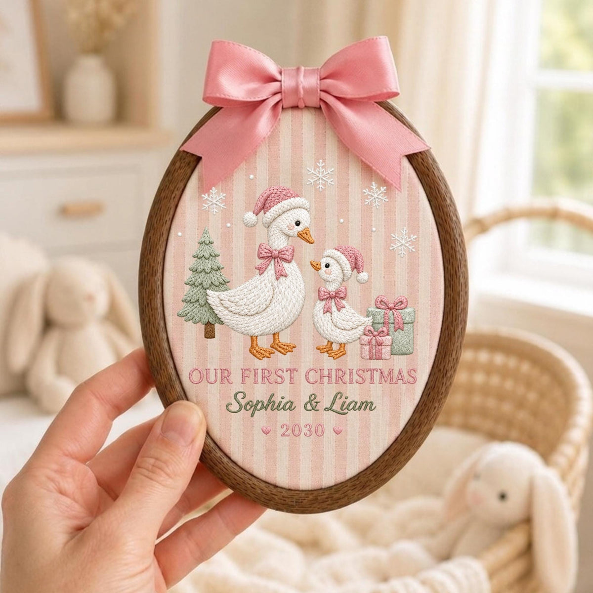 May include: An oval wooden embroidery hoop featuring a pink and white striped fabric with an embroidered design of two geese wearing pink Santa hats, a green tree, and gifts. The text reads "OUR FIRST CHRISTMAS Sophia &amp; Liam 2030" with a pink bow.