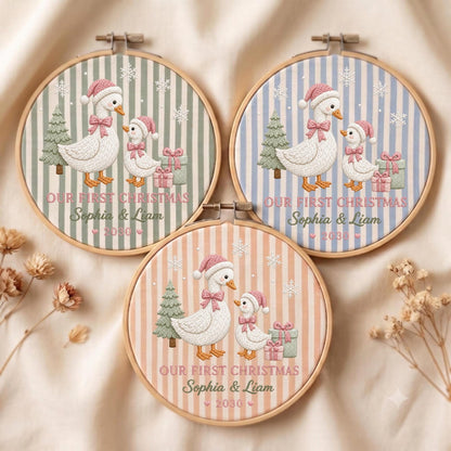 May include: Three circular wooden embroidery hoops featuring fabric with vertical stripes in sage green, light blue, and peach. Each displays embroidered white geese wearing pink hats and bows, a green tree, gifts, snowflakes, and the text OUR FIRST CHRISTMAS Sophia &amp; Liam 2030.