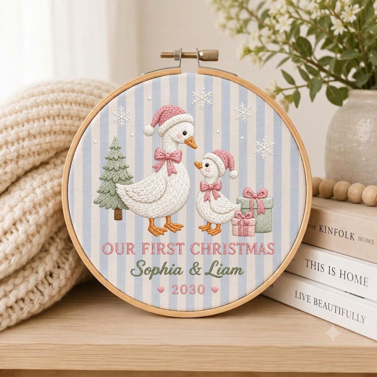 May include: A circular embroidery hoop featuring a light blue and white striped fabric background. The design includes two white geese wearing pink Santa hats and bows, a green Christmas tree, gifts, and snowflakes. Text reads: OUR FIRST CHRISTMAS Sophia &amp; Liam 2030.
