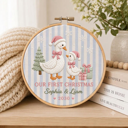 May include: A circular embroidery hoop featuring a light blue and white striped fabric background. The design includes two white geese wearing pink Santa hats and bows, a green Christmas tree, gifts, and snowflakes. Text reads: OUR FIRST CHRISTMAS Sophia &amp; Liam 2030.