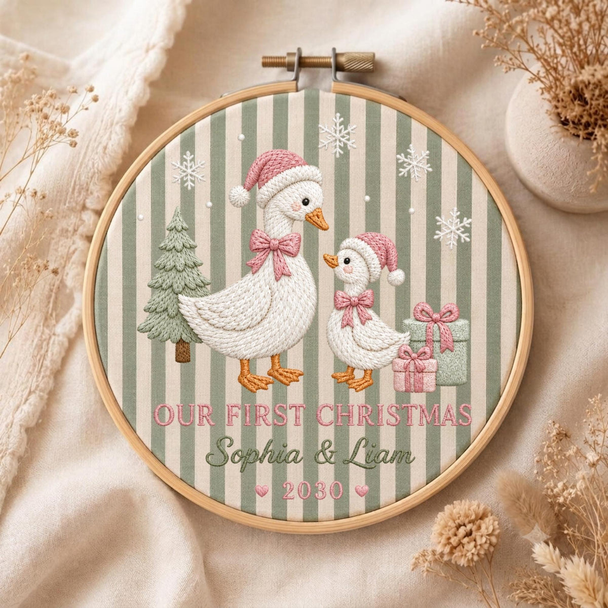 May include: A circular embroidery hoop featuring a green and white striped fabric background. The design depicts two white geese wearing pink Santa hats and bows, a green Christmas tree, and two gift boxes. Text reads: OUR FIRST CHRISTMAS Sophia &amp; Liam 2030.