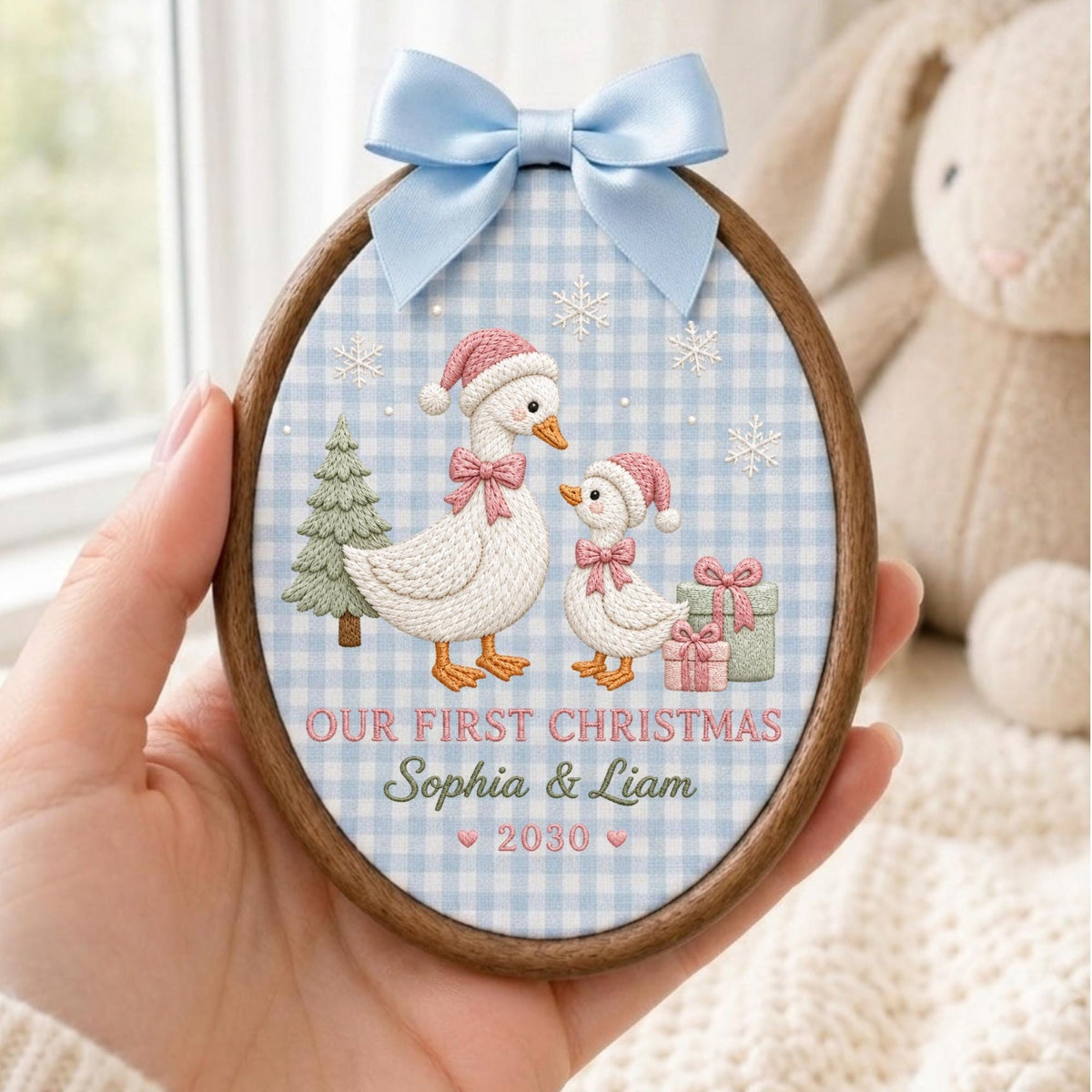 May include: An oval wooden embroidery hoop ornament featuring a light blue gingham fabric background. It displays two embroidered geese wearing pink hats and bows, a green tree, gifts, and snowflakes. Text reads: OUR FIRST CHRISTMAS Sophia &amp; Liam 2030.