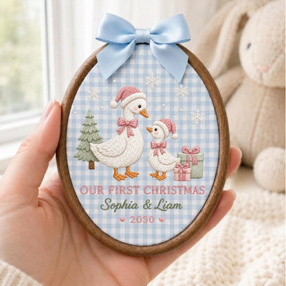 May include: An oval wooden embroidery hoop ornament featuring a light blue gingham fabric background. It displays two embroidered geese wearing pink hats and bows, a green tree, gifts, and snowflakes. Text reads: OUR FIRST CHRISTMAS Sophia &amp; Liam 2030.