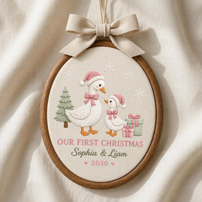 May include: An oval wooden embroidery hoop containing fabric with an embroidered scene of two geese wearing pink hats and bows, a green tree, and gifts. The text reads "OUR FIRST CHRISTMAS", "Sophia &amp; Liam", and "2030". A beige ribbon bow is attached.