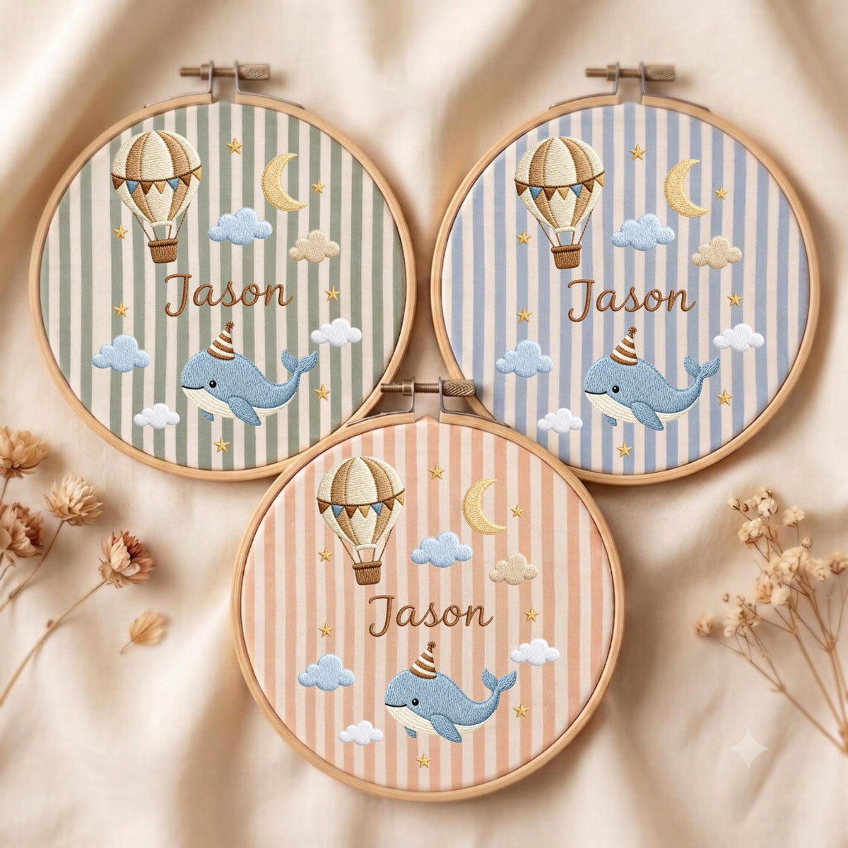 May include: Three circular wooden embroidery hoops featuring fabric with vertical stripes in sage green, light blue, and peach. Each hoop displays embroidered designs of a hot air balloon, a crescent moon, stars, clouds, and a whale wearing a party hat, with the name Jason stitched in brown thread.