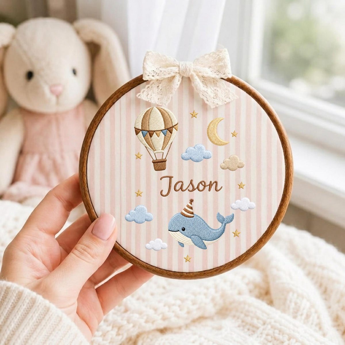 May include: A circular wooden embroidery hoop featuring a light pink and white striped fabric background. The design includes an embroidered hot air balloon, a crescent moon, clouds, and a blue whale wearing a party hat, with the name Jason.