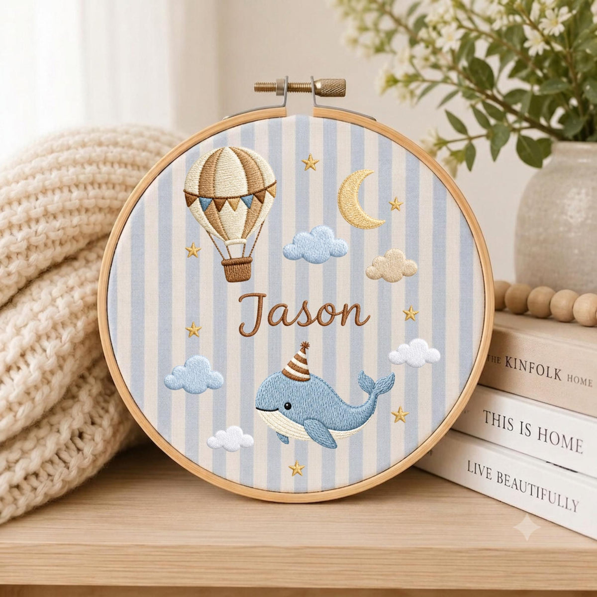 May include: A circular embroidery hoop featuring a light blue and white striped fabric background. The design includes a tan hot air balloon, a yellow crescent moon, white and blue clouds, gold stars, and a blue whale wearing a striped party hat.