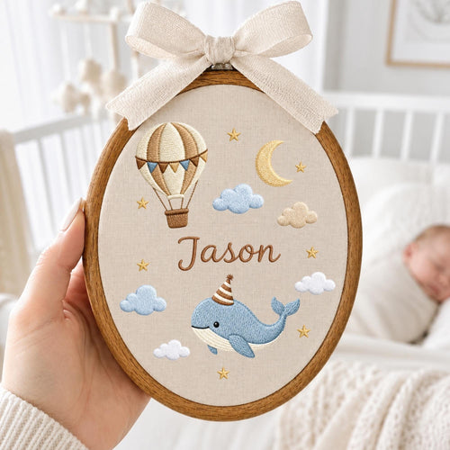 May include: An oval wooden embroidery hoop containing beige fabric with a stitched design of a hot air balloon, a crescent moon, clouds, and a blue whale wearing a party hat. The name Jason is embroidered in brown. A beige ribbon bow is at the top.