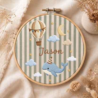 May include: A round wooden embroidery hoop containing fabric with vertical sage green and cream stripes. The embroidery features a brown hot air balloon, a yellow crescent moon, blue and white clouds, yellow stars, and a blue whale wearing a party hat, with the name Jason stitched in brown thread.