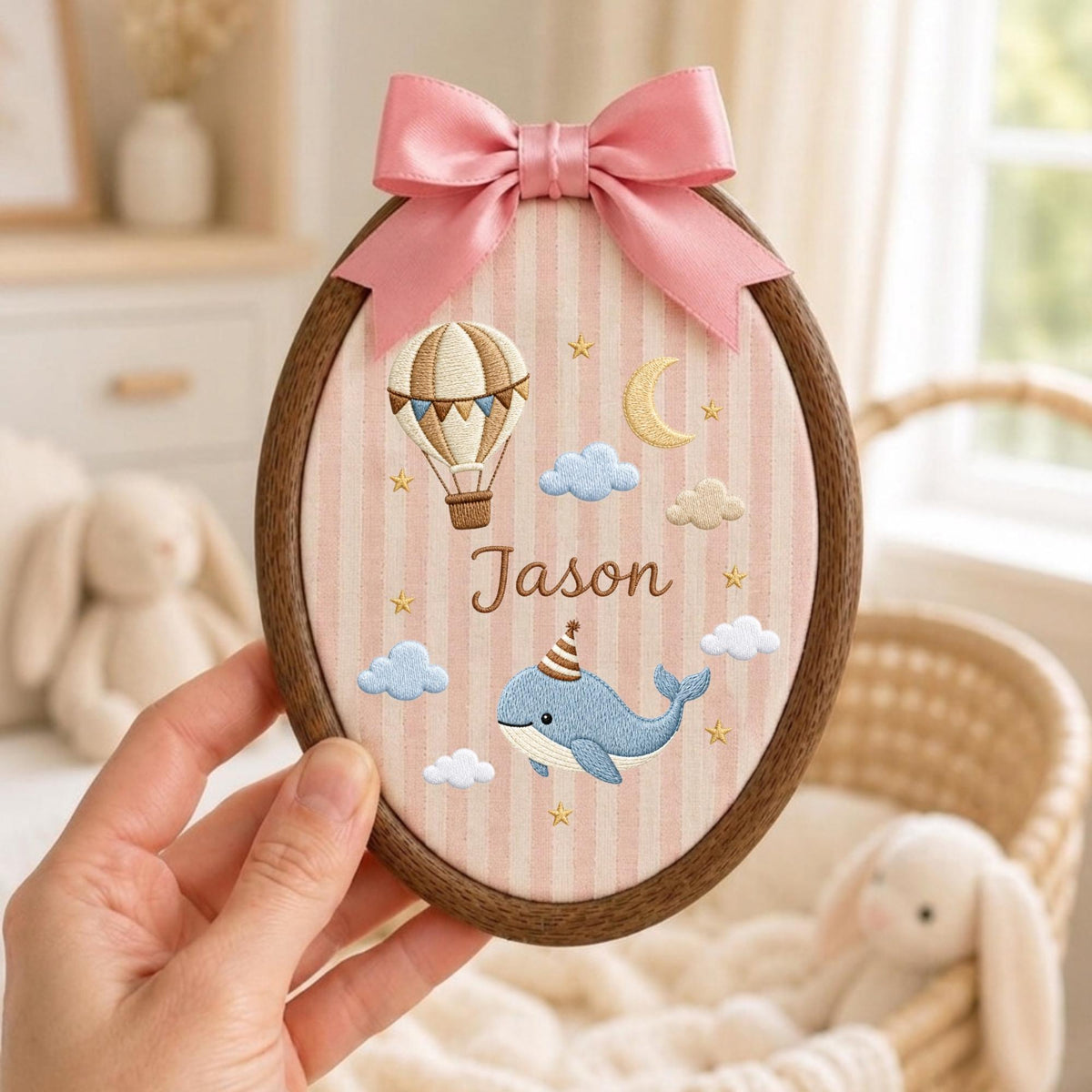 May include: An oval wooden embroidery hoop featuring a light pink and white striped fabric background. The fabric is embroidered with a brown hot air balloon, a yellow crescent moon, and a blue whale wearing a party hat, with the name Jason.