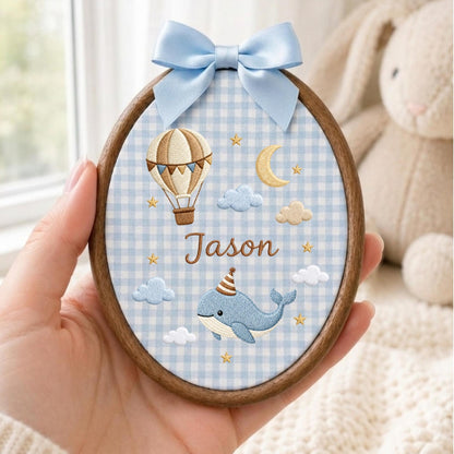 May include: An oval wooden embroidery hoop featuring light blue and white gingham fabric. The design includes an embroidered hot air balloon, a crescent moon, clouds, stars, and a blue whale wearing a party hat. The name Jason is stitched in brown.