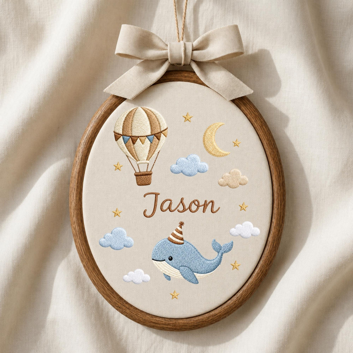 May include: An oval wooden embroidery hoop containing cream fabric with a beige bow at the top. The embroidery features a hot air balloon, a crescent moon, clouds, stars, and a blue whale wearing a party hat, with the name Jason in brown.