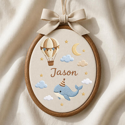May include: An oval wooden embroidery hoop containing cream fabric with a beige bow at the top. The embroidery features a hot air balloon, a crescent moon, clouds, stars, and a blue whale wearing a party hat, with the name Jason in brown.