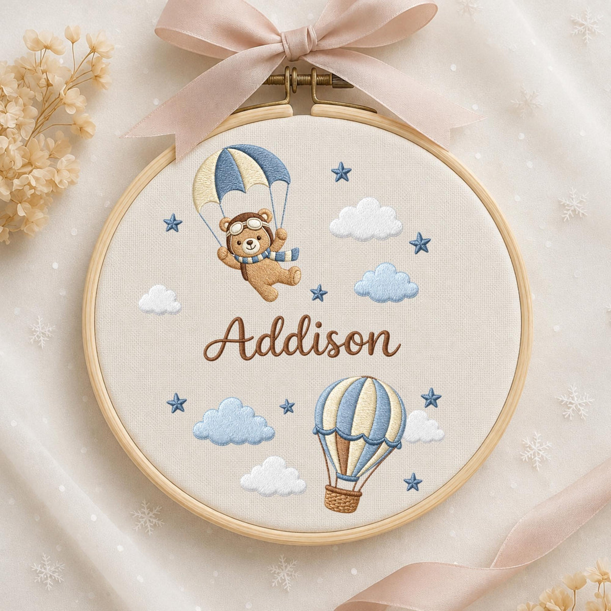 May include: A round wooden embroidery hoop containing cream-colored fabric with an embroidered design of a brown bear in a pilot hat with a parachute, a hot air balloon, clouds, and stars. The name Addison is stitched in brown cursive text.
