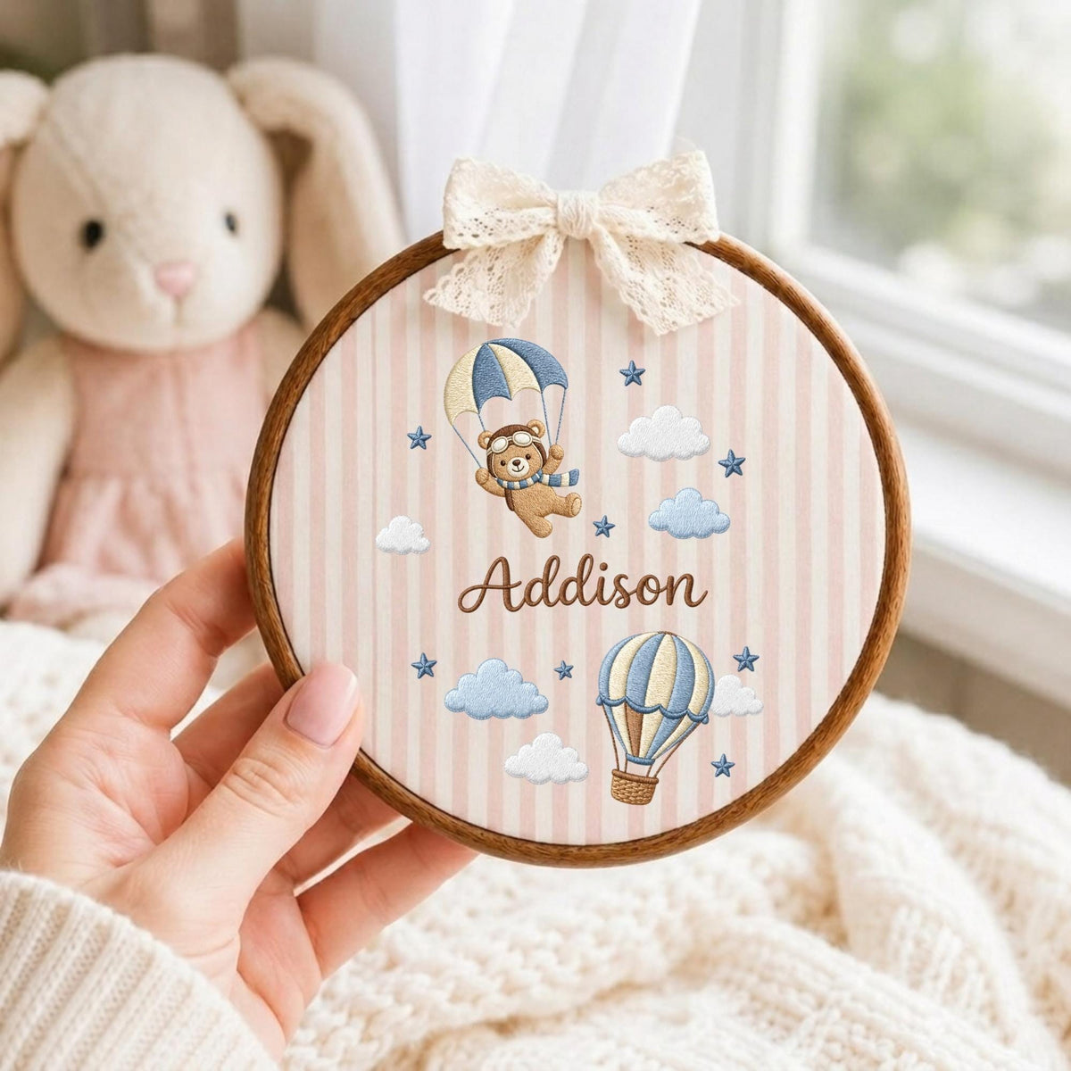 May include: A circular wooden embroidery hoop containing fabric with vertical pink and white stripes. The fabric features an embroidered brown bear in a parachute, a hot air balloon, clouds, stars, and the name Addison in brown script, topped with a lace bow.