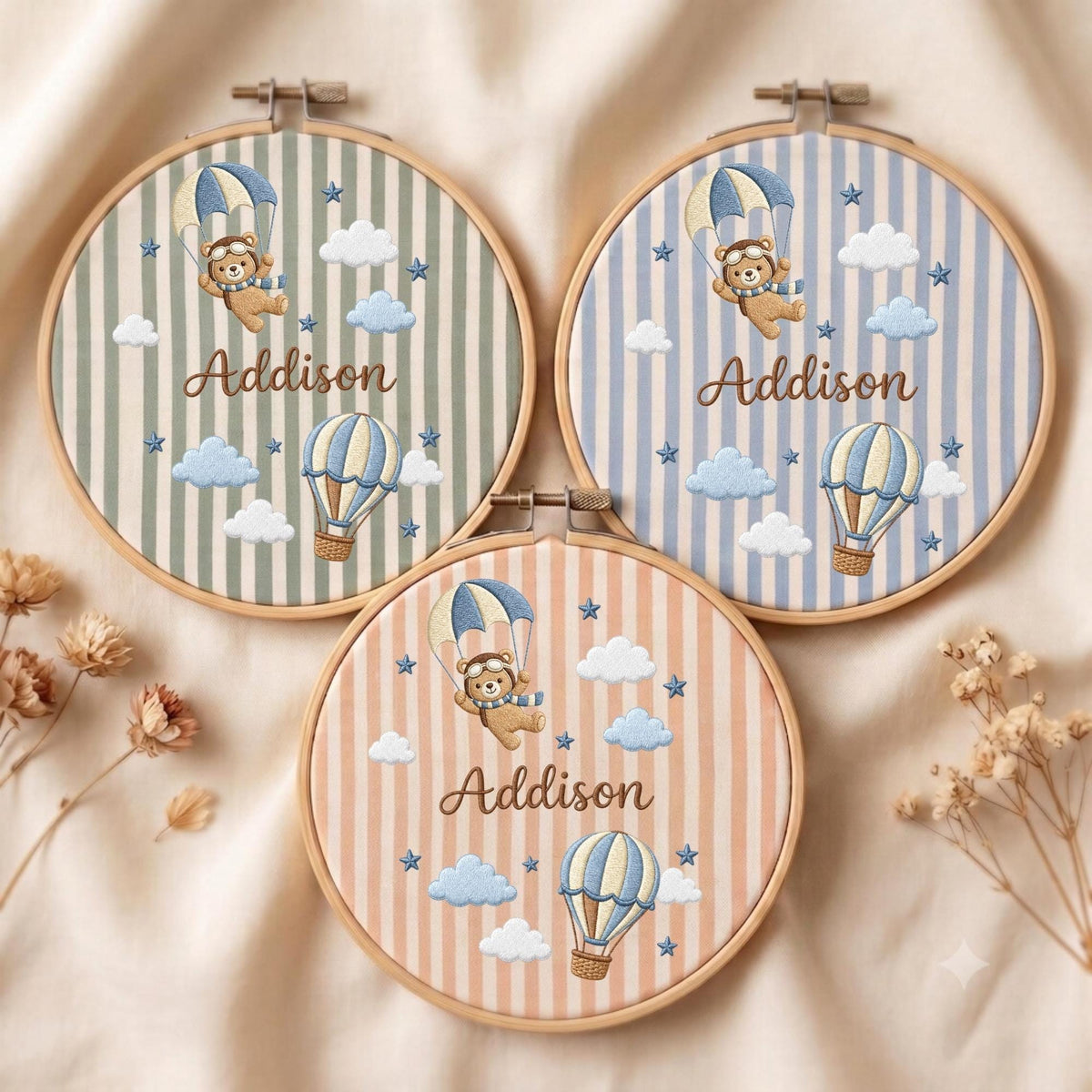 May include: Three circular wooden embroidery hoops featuring fabric with vertical stripes in sage green, light blue, and peach. Each hoop displays an embroidered teddy bear in a parachute, a hot air balloon, clouds, stars, and the name Addison.