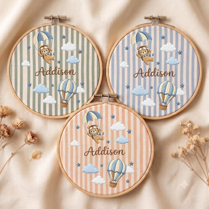 May include: Three circular wooden embroidery hoops featuring fabric with vertical stripes in sage green, light blue, and peach. Each hoop displays an embroidered teddy bear in a parachute, a hot air balloon, clouds, stars, and the name Addison.