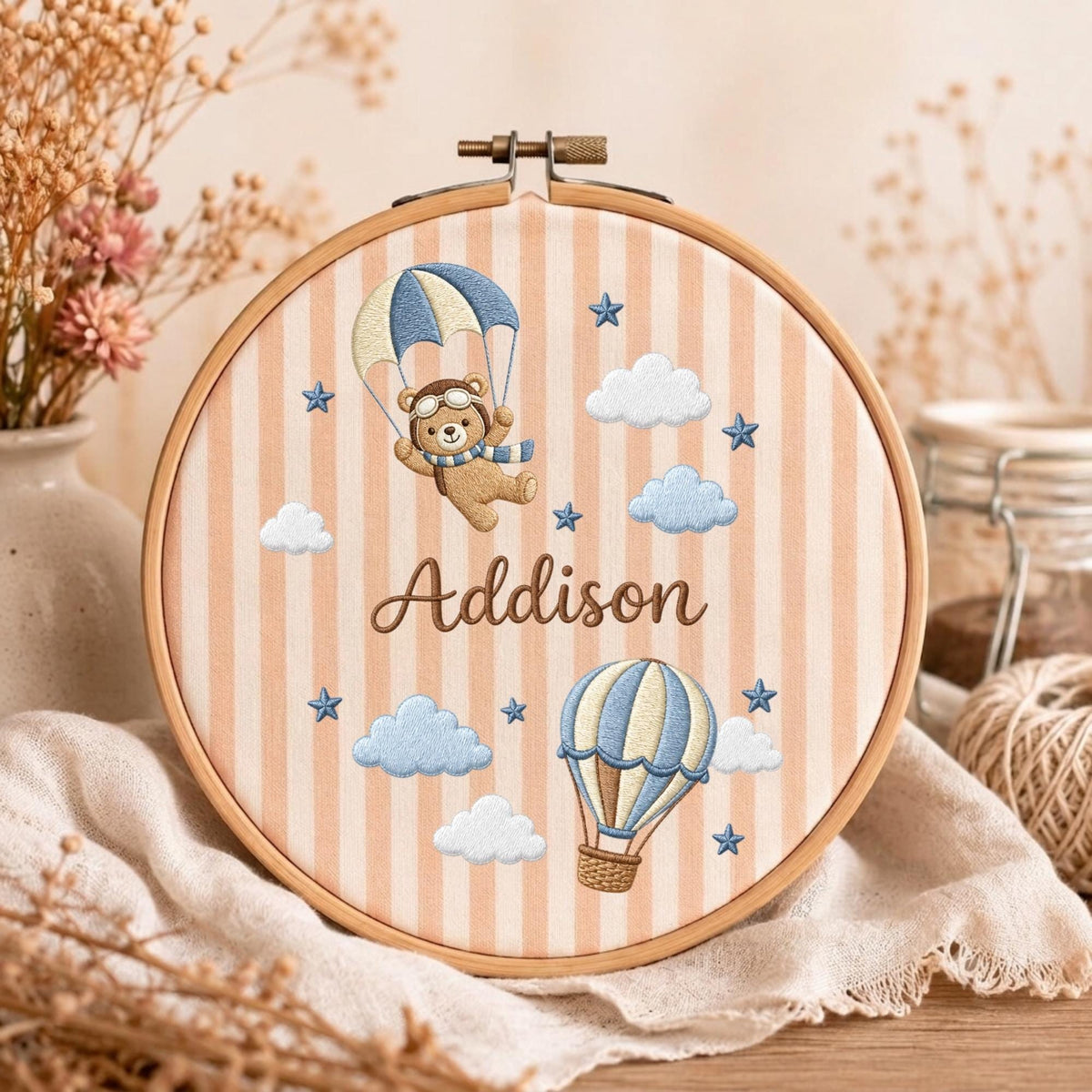 May include: A circular wooden embroidery hoop containing fabric with vertical peach and white stripes. The embroidery features a brown bear in a pilot hat with a blue hot air balloon, a separate hot air balloon, clouds, stars, and the name Addison.