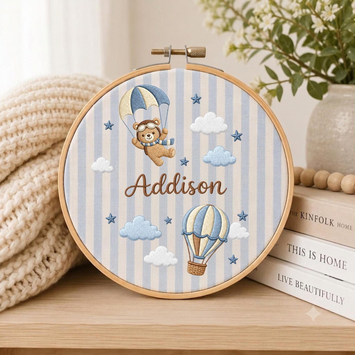 May include: A circular wooden embroidery hoop containing fabric with light blue and white vertical stripes. The fabric features an embroidered brown bear in a parachute, a hot air balloon, clouds, stars, and the name Addison in brown script.