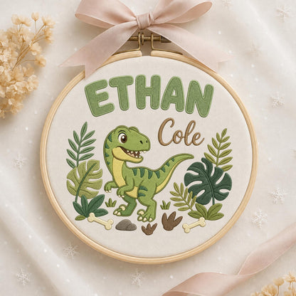 May include: A circular wooden embroidery hoop containing white fabric with a stitched green dinosaur, tropical leaves, and bones. The text ETHAN is embroidered in large green letters above the dinosaur, with the name Cole in brown script.