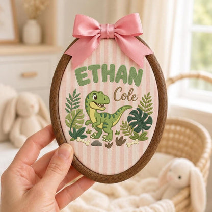 May include: An oval embroidery hoop wall hanging featuring a light pink and white striped fabric background. The design includes the embroidered name Ethan Cole in green and brown, a green dinosaur, and tropical leaves with a pink satin bow on top.