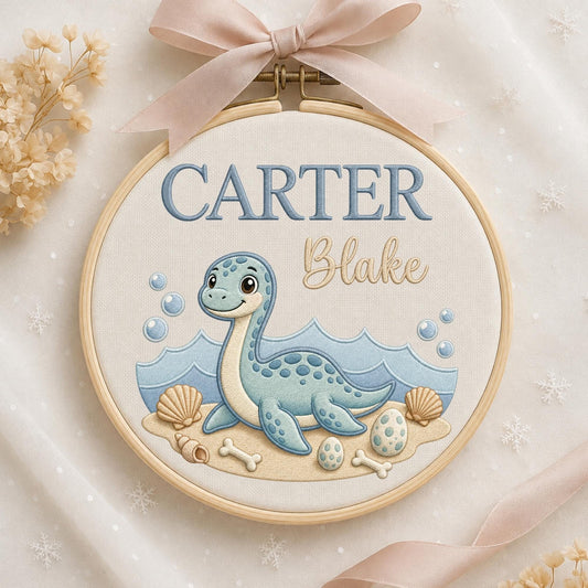 May include: A circular wooden embroidery hoop containing fabric with an embroidered blue dinosaur, waves, seashells, and eggs. The text reads CARTER in blue block letters and Blake in tan script. A light pink ribbon is tied at the top of the hoop.