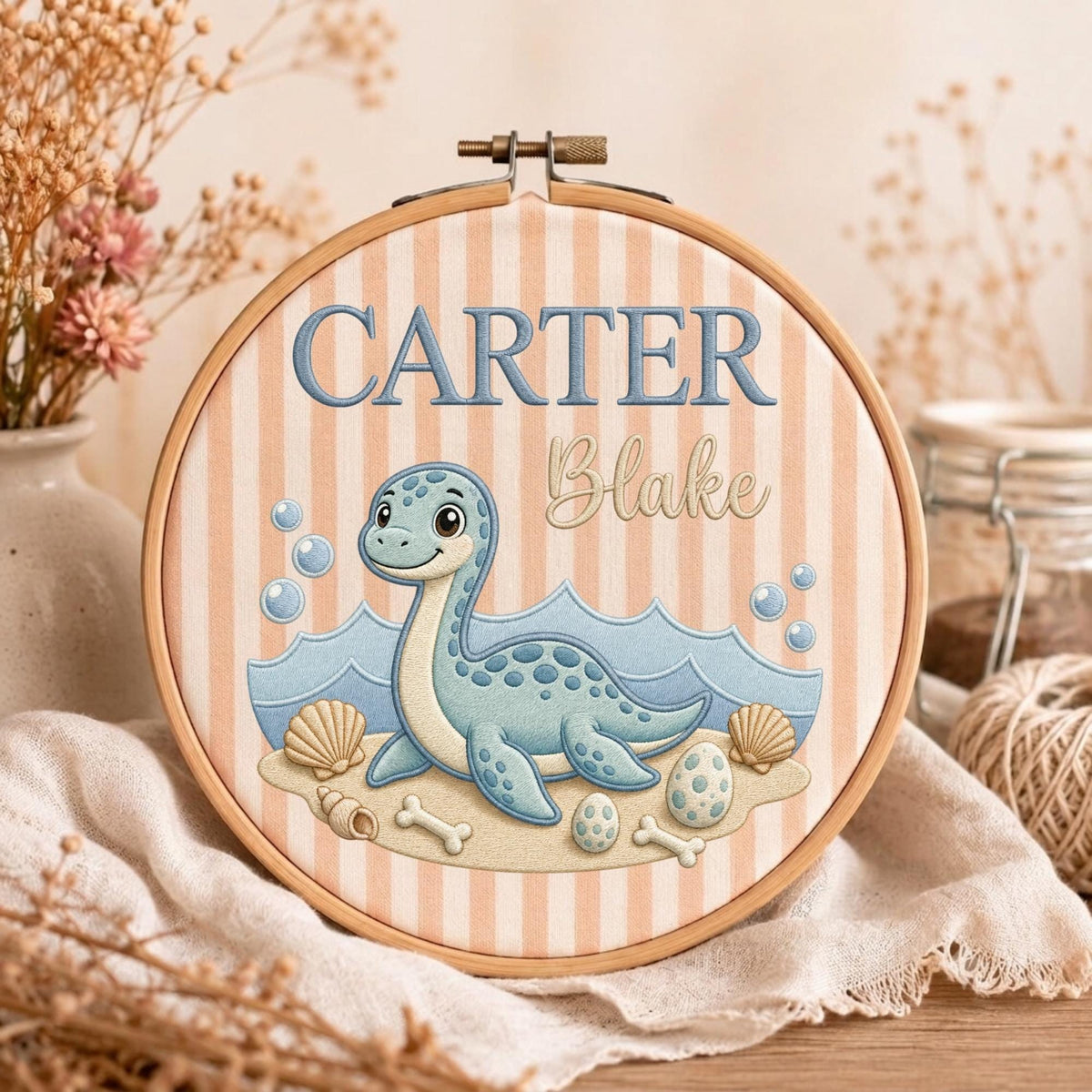 May include: A circular wooden embroidery hoop framing fabric with vertical peach and white stripes. The design features the embroidered text CARTER Blake above a blue dinosaur on a sandy beach with waves, seashells, eggs, and bones.