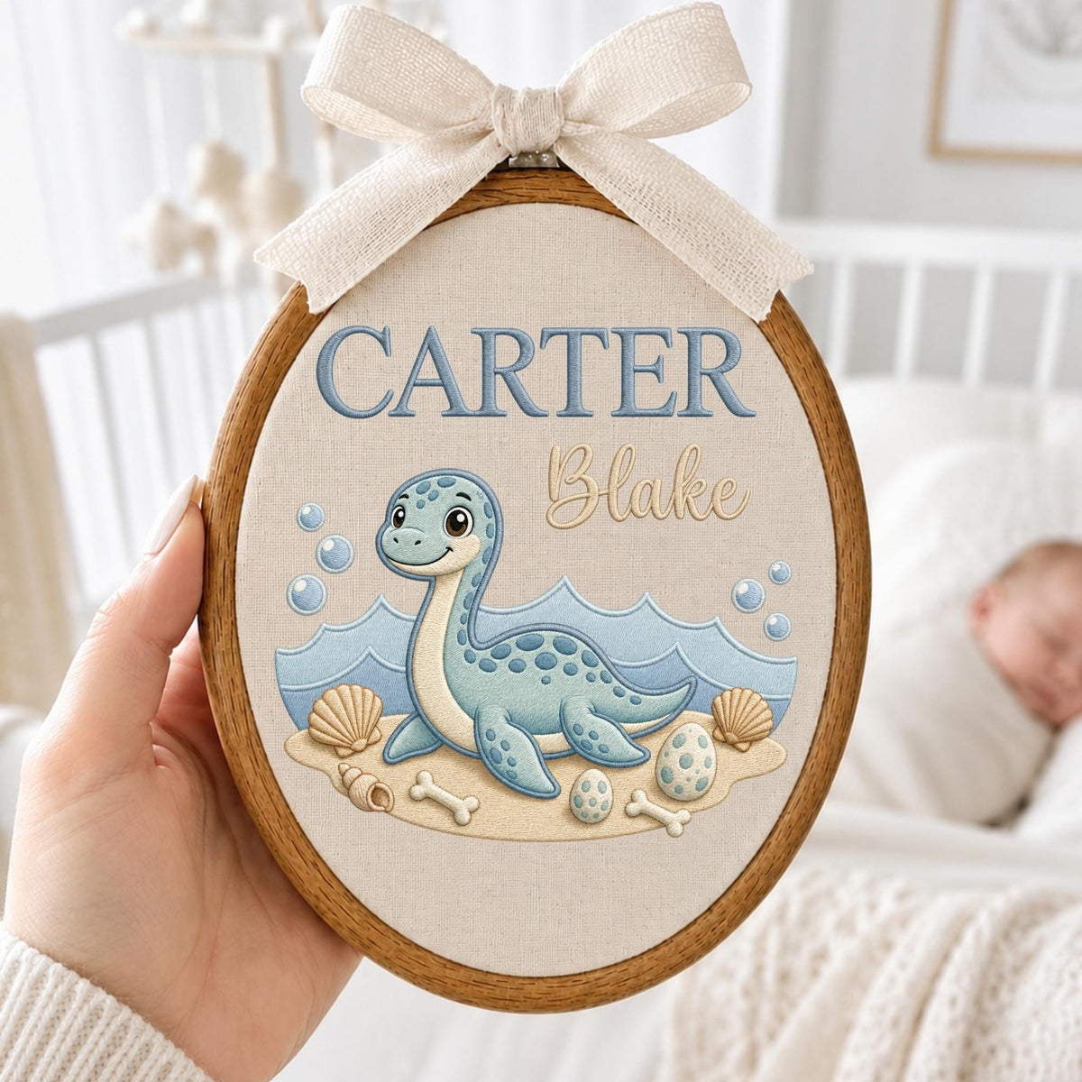 May include: An oval wooden embroidery hoop containing a fabric panel with the embroidered text CARTER Blake. The design features a light blue aquatic creature on a sandy beach with seashells, eggs, and bones, topped with a beige fabric bow.