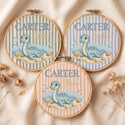 May include: Three circular embroidery hoops featuring a light blue cartoon dinosaur on a sandy beach with seashells and bubbles. Each hoop has vertical stripes in sage green, light blue, or peach, with the text CARTER Blake embroidered on top.