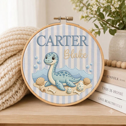 May include: A circular embroidery hoop featuring a light blue and white striped fabric background. The design includes a light blue dinosaur, seashells, eggs, and bones on a sandy base. The text reads CARTER in blue and Blake in gold script.