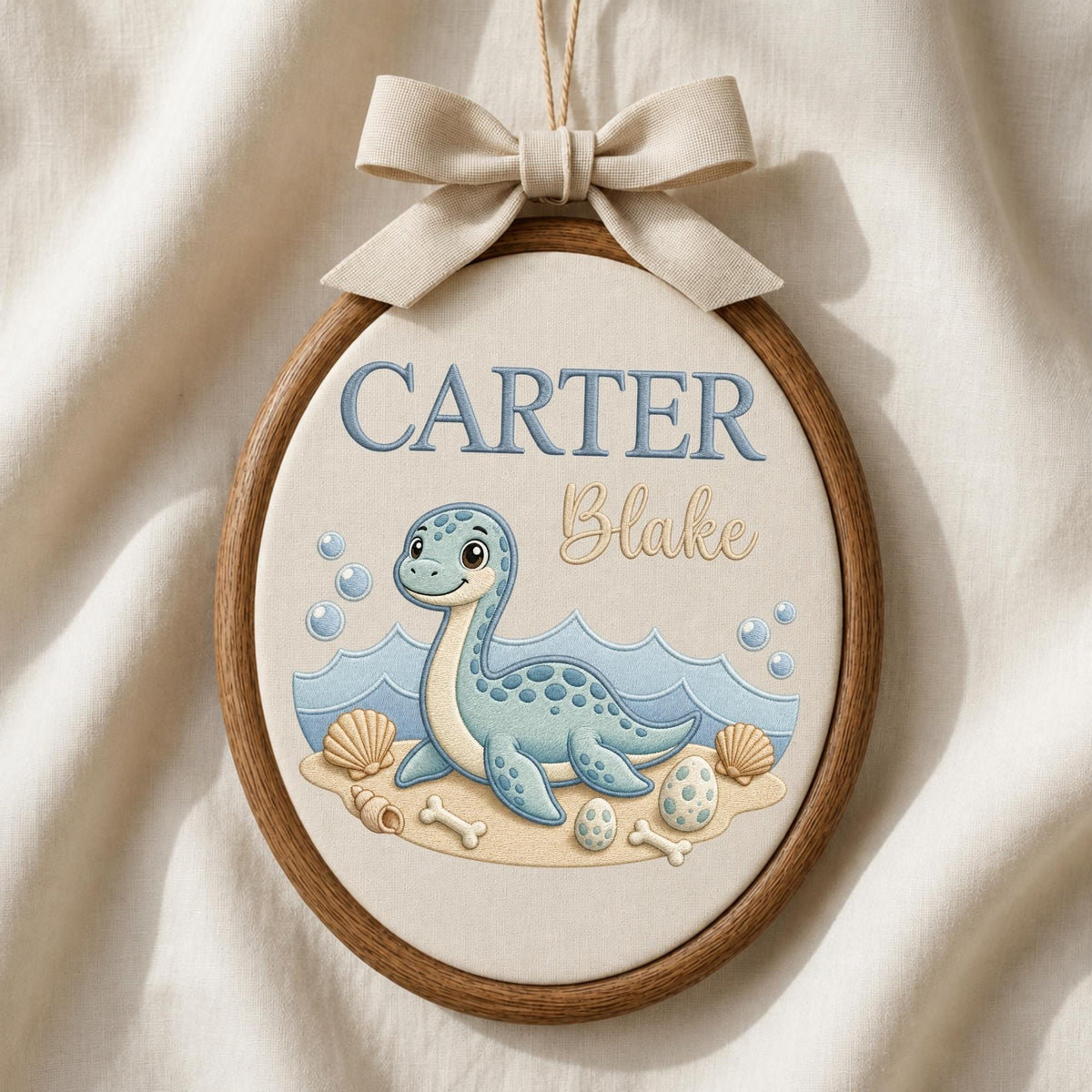 May include: An oval embroidery hoop wall hanging featuring a light blue dinosaur illustration on cream fabric. The text reads CARTER in blue and Blake in tan script. The design includes blue waves, bubbles, seashells, eggs, and bones with a beige bow.