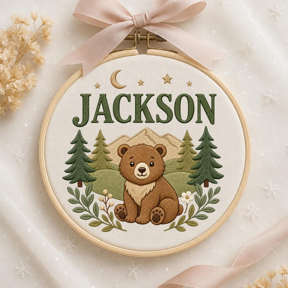 May include: A circular wooden embroidery hoop containing white fabric with a stitched design of a brown bear, green pine trees, tan mountains, and a crescent moon with stars. The name JACKSON is embroidered in green above the bear.