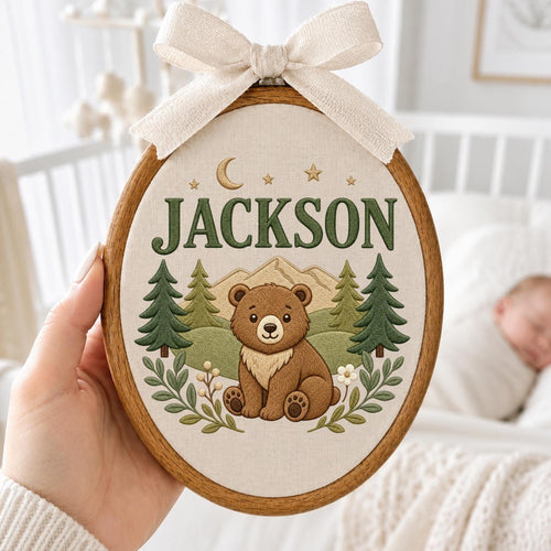 May include: An oval wooden embroidery hoop containing fabric with the embroidered name JACKSON in dark green letters. The design features a brown bear sitting in front of green pine trees and tan mountains, with a crescent moon and stars above.