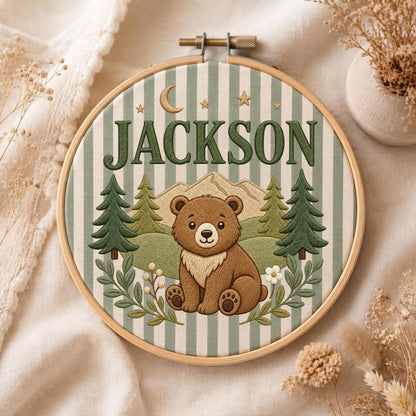 May include: A circular embroidery hoop featuring a light green and white striped fabric background. The design includes a brown bear sitting in front of embroidered mountains and pine trees, with the name JACKSON and a crescent moon and stars.