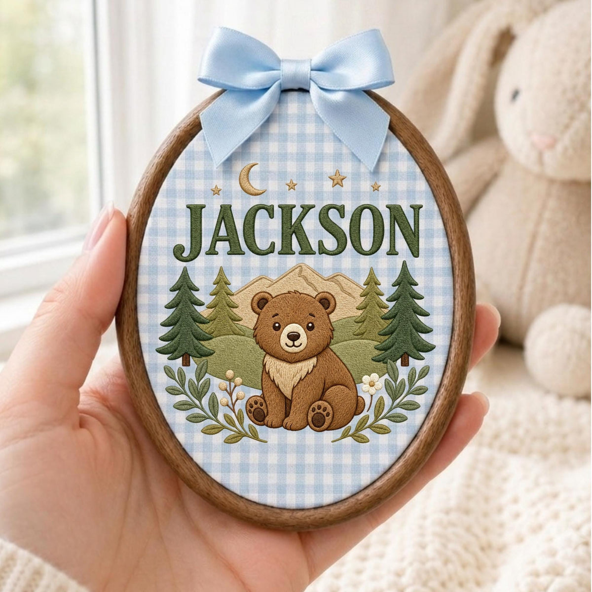 May include: An oval wooden embroidery hoop frame containing fabric with a light blue gingham pattern. The design features a brown bear, green pine trees, mountains, stars, a crescent moon, and the name JACKSON in dark green capital letters.