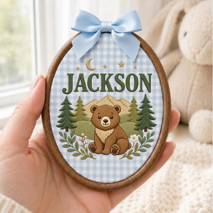 May include: An oval wooden embroidery hoop frame containing fabric with a light blue gingham pattern. The design features a brown bear, green pine trees, mountains, stars, a crescent moon, and the name JACKSON in dark green capital letters.