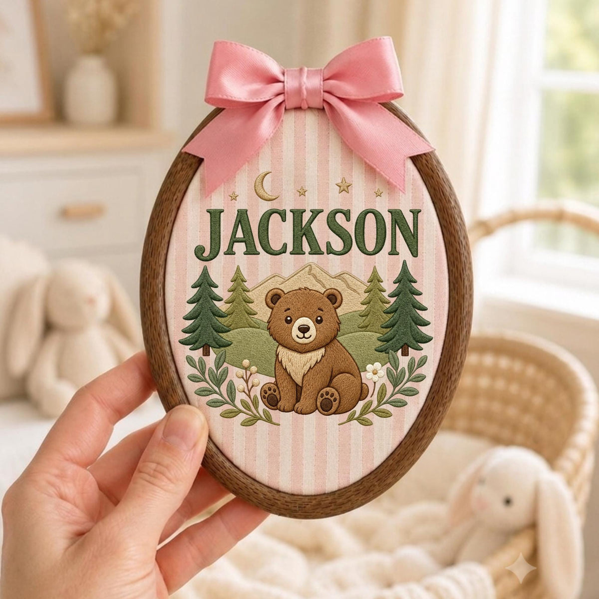 May include: An oval wooden embroidery hoop frame containing fabric with a pink and white striped pattern. The fabric features an embroidered brown bear, green pine trees, a crescent moon, stars, and the name JACKSON in dark green letters.