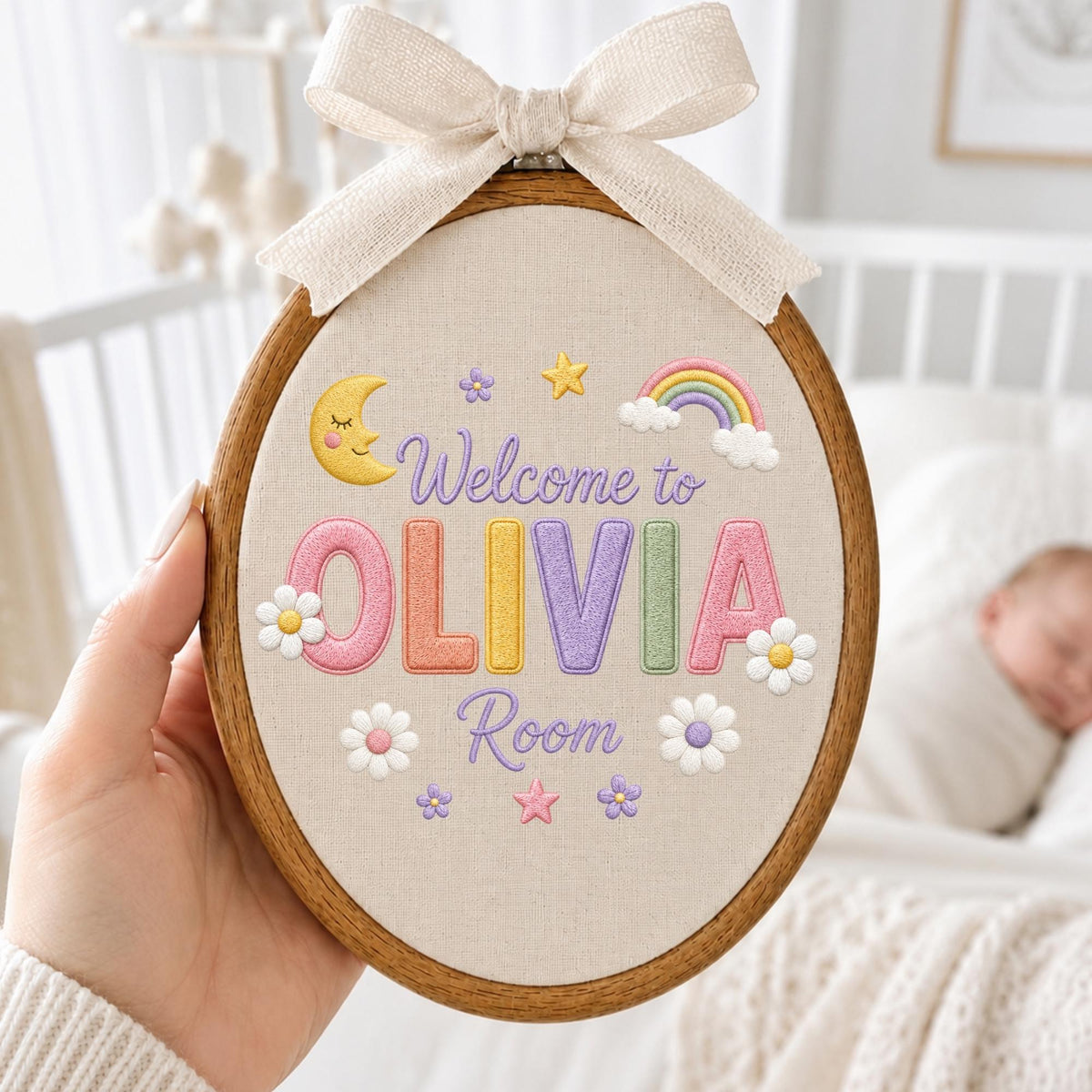 May include: An oval wooden embroidery hoop containing beige fabric with the embroidered text Welcome to OLIVIA Room. The design features a yellow crescent moon, a rainbow, stars, small flowers, and a large beige fabric bow at the top.