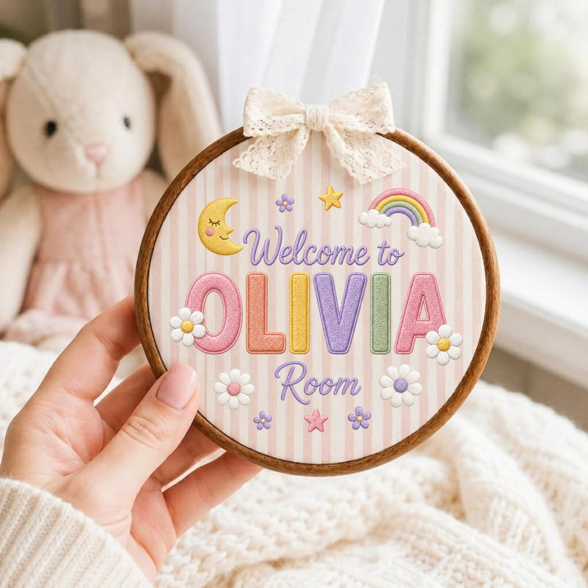 May include: A circular wooden embroidery hoop containing fabric with vertical light pink stripes. The fabric features embroidered text reading Welcome to OLIVIA Room, surrounded by a yellow crescent moon, a rainbow, stars, and white flowers.