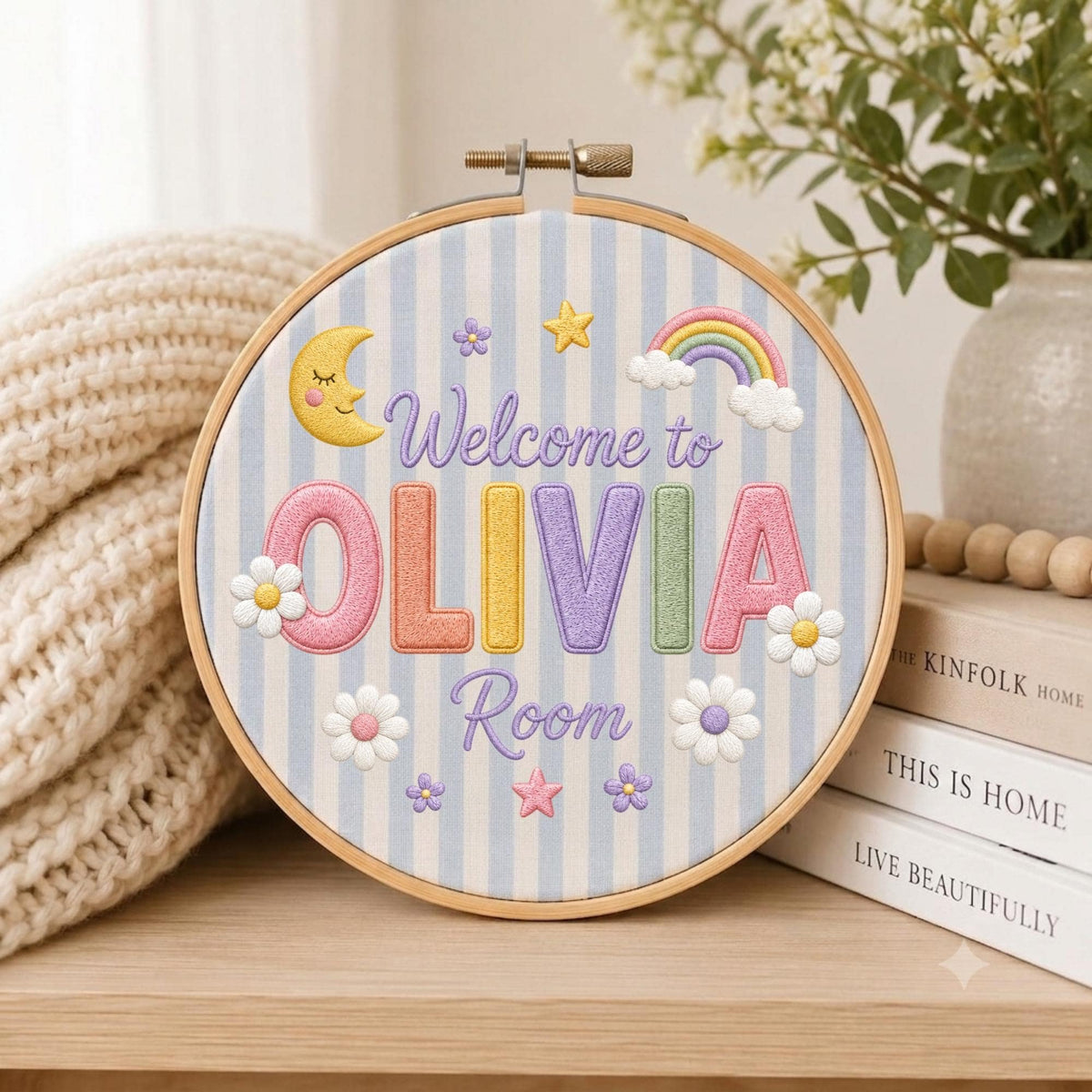 May include: A circular embroidery hoop featuring a light blue and white striped fabric background. The text reads Welcome to OLIVIA Room in colorful embroidered letters. Decorative elements include a yellow crescent moon, a rainbow, and flowers.