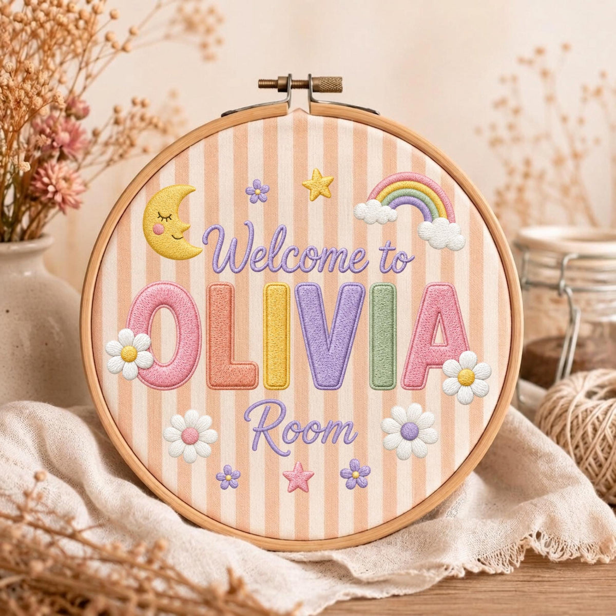 May include: A circular wooden embroidery hoop containing fabric with vertical beige stripes. The embroidery features the text "Welcome to OLIVIA Room" in purple, surrounded by a yellow crescent moon, a rainbow, and several small flowers and stars.