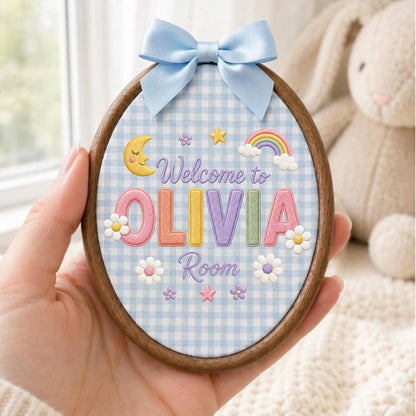 May include: An oval embroidery hoop with a light blue gingham fabric background. The text reads Welcome to OLIVIA Room in colorful letters. Embroidered motifs include a yellow crescent moon, a rainbow, stars, and white flowers. A light blue satin bow is attached to the top.