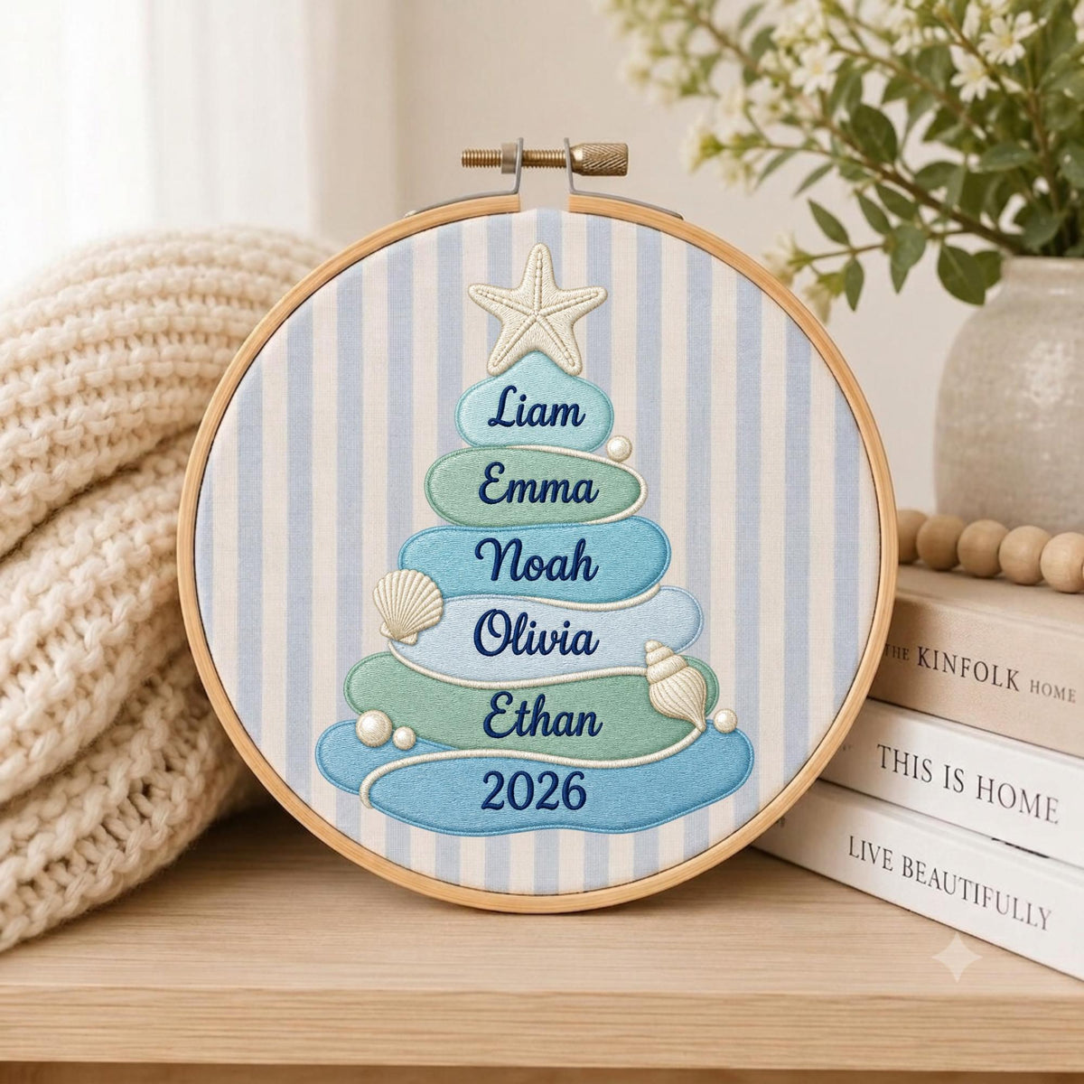 May include: A circular embroidery hoop featuring a light blue and white striped fabric background. Centered is a stylized Christmas tree made of stacked, rounded blue and green shapes, topped with a starfish, decorated with seashells, and embroidered with the names Liam, Emma, Noah, Olivia, Ethan, and the year 2026.