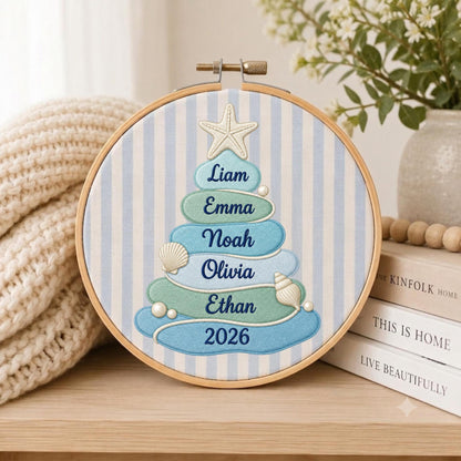 May include: A circular embroidery hoop featuring a light blue and white striped fabric background. Centered is a stylized Christmas tree made of stacked, rounded blue and green shapes, topped with a starfish, decorated with seashells, and embroidered with the names Liam, Emma, Noah, Olivia, Ethan, and the year 2026.