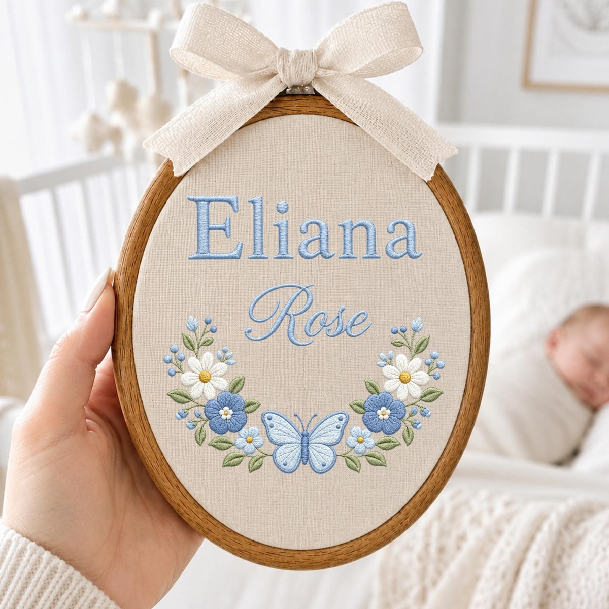 May include: An oval wooden embroidery hoop containing beige fabric with the embroidered blue text Eliana Rose. Below the text is a floral wreath featuring white and blue flowers, green leaves, and a central light blue butterfly, topped with a beige bow.