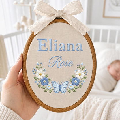 May include: An oval wooden embroidery hoop containing beige fabric with the embroidered blue text Eliana Rose. Below the text is a floral wreath featuring white and blue flowers, green leaves, and a central light blue butterfly, topped with a beige bow.