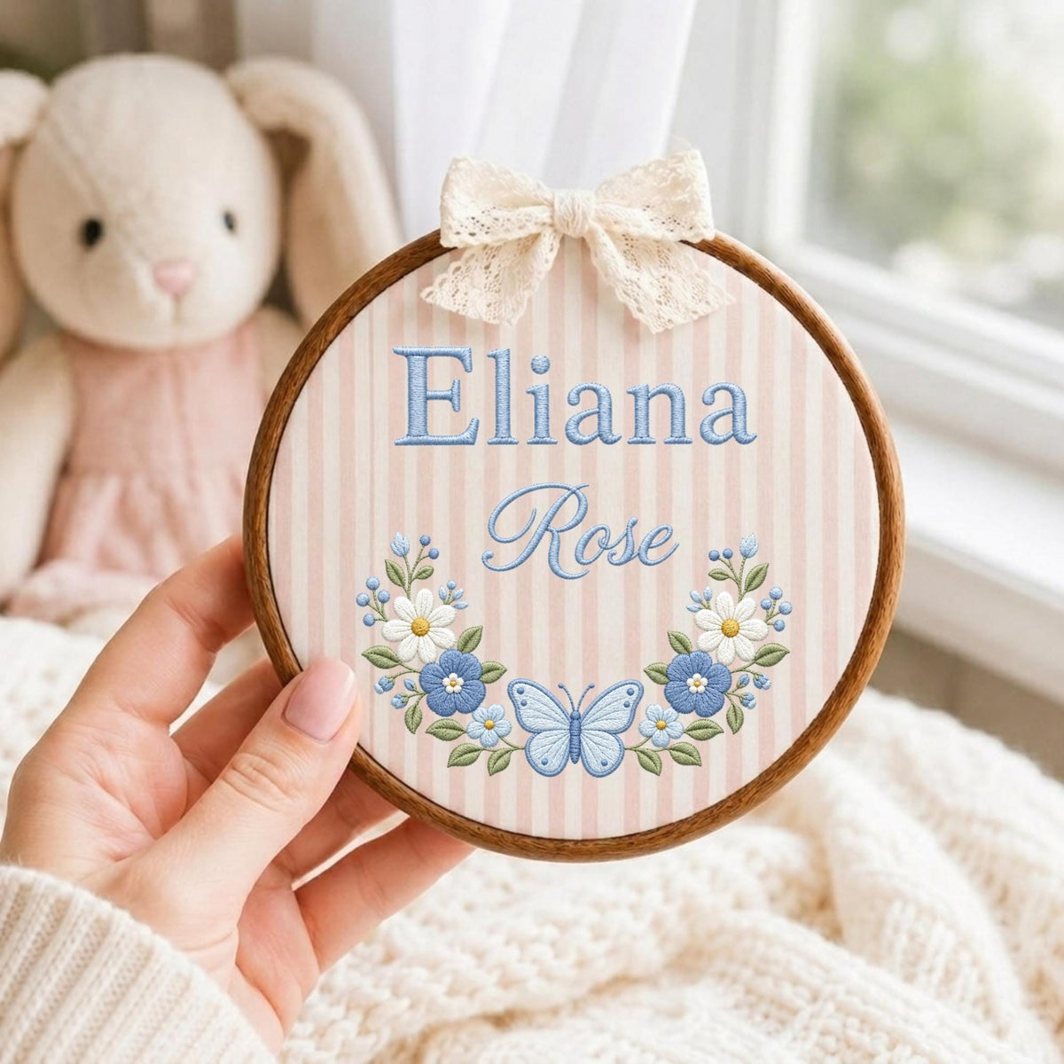 May include: A circular wooden embroidery hoop featuring a light pink and white striped fabric background. The text Eliana Rose is embroidered in blue, centered above a floral wreath with a light blue butterfly and white and blue flowers.