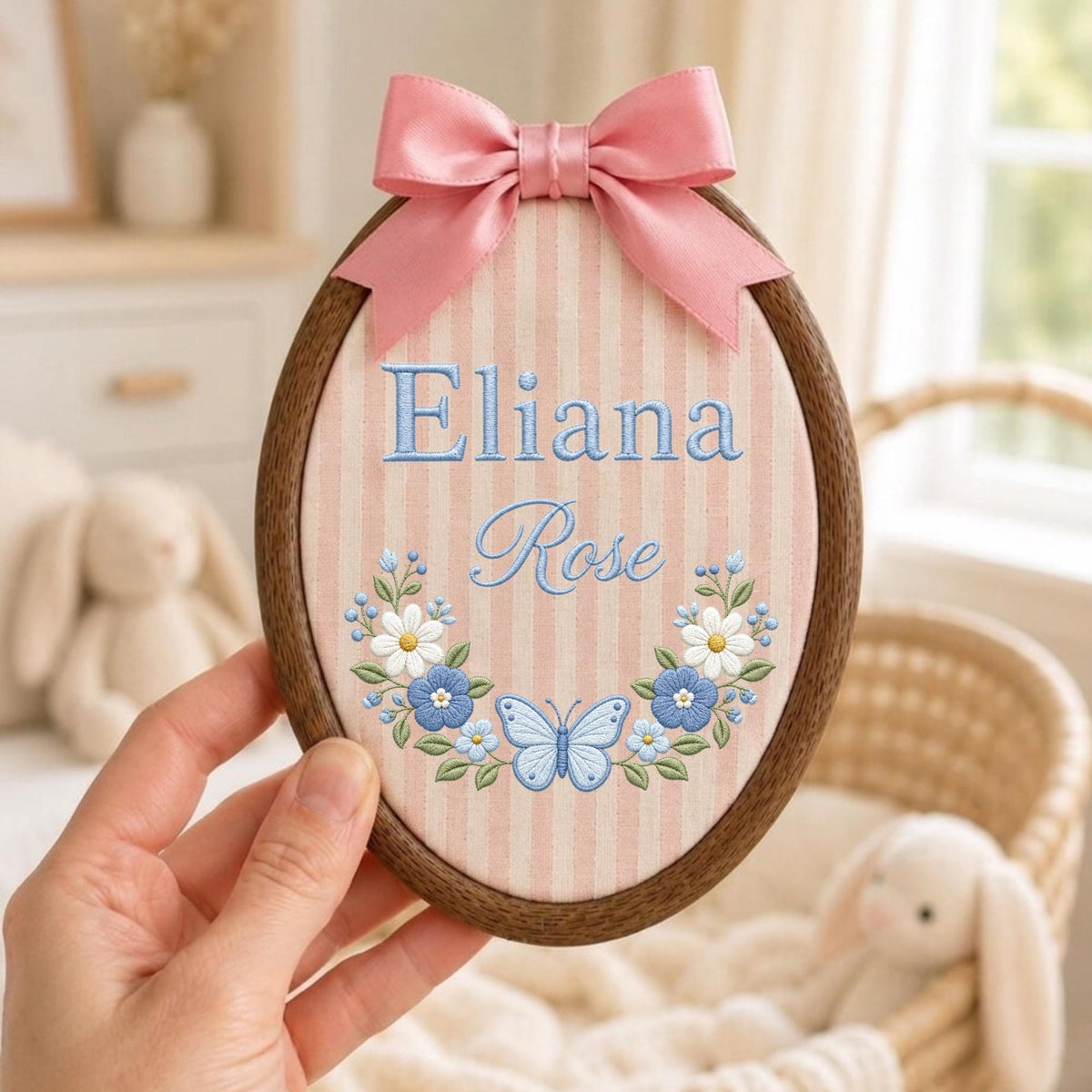 May include: An oval wooden embroidery hoop containing fabric with vertical pink and white stripes. The fabric features the embroidered name Eliana Rose in blue, accented by a floral wreath and a light blue butterfly, topped with a pink satin bow.