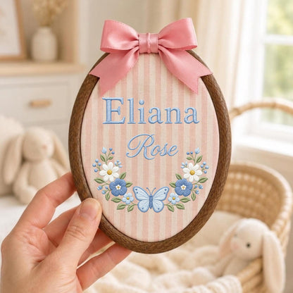 May include: An oval wooden embroidery hoop containing fabric with vertical pink and white stripes. The fabric features the embroidered name Eliana Rose in blue, accented by a floral wreath and a light blue butterfly, topped with a pink satin bow.